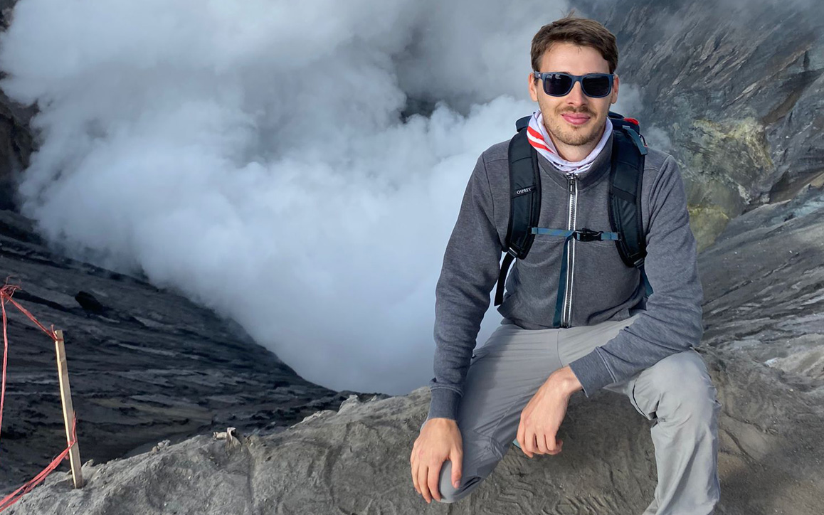 Constantin Haider sitzt mit Rucksack und Sonnenbrille auf einem Felsen vor einer dampfenden Vulkanlandschaft.
