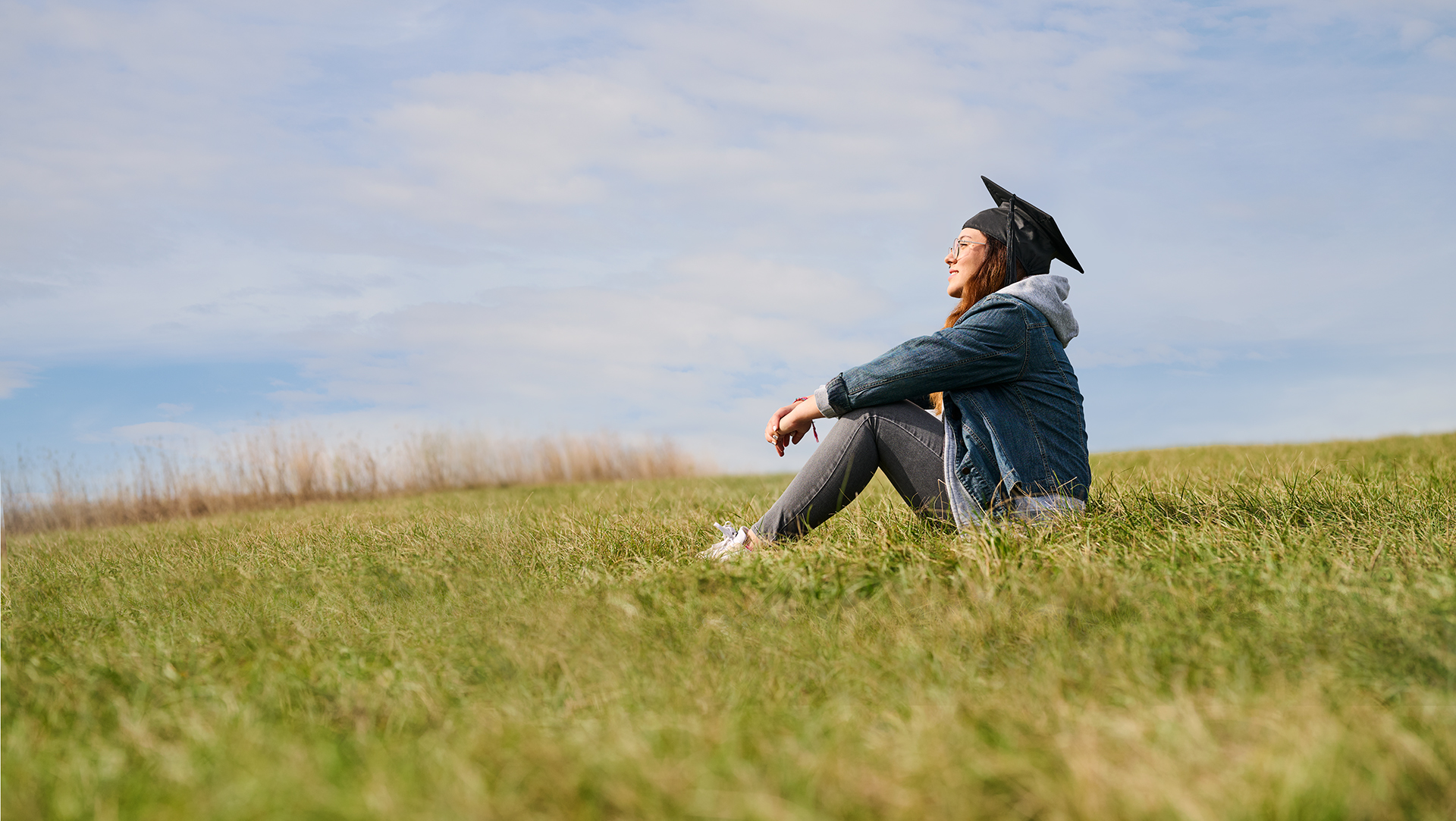 A student with a graduate hat sits relaxed on a meadow and looks up at the sky - symbolising individual support, personal development and future prospects at University of Applied Sciences Burgenland.